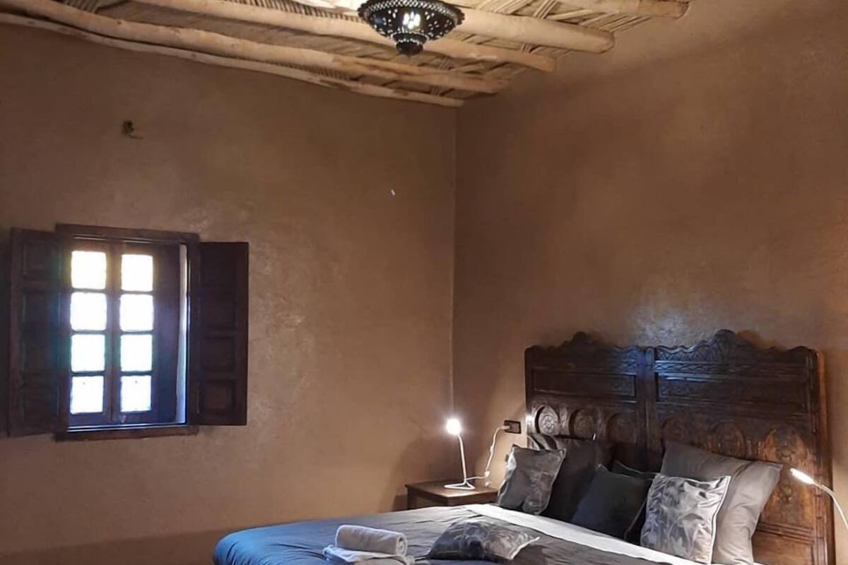 A bedroom with a bed, a nightstand, a lamp, a window with shutters, and a ceiling with a woven pattern.