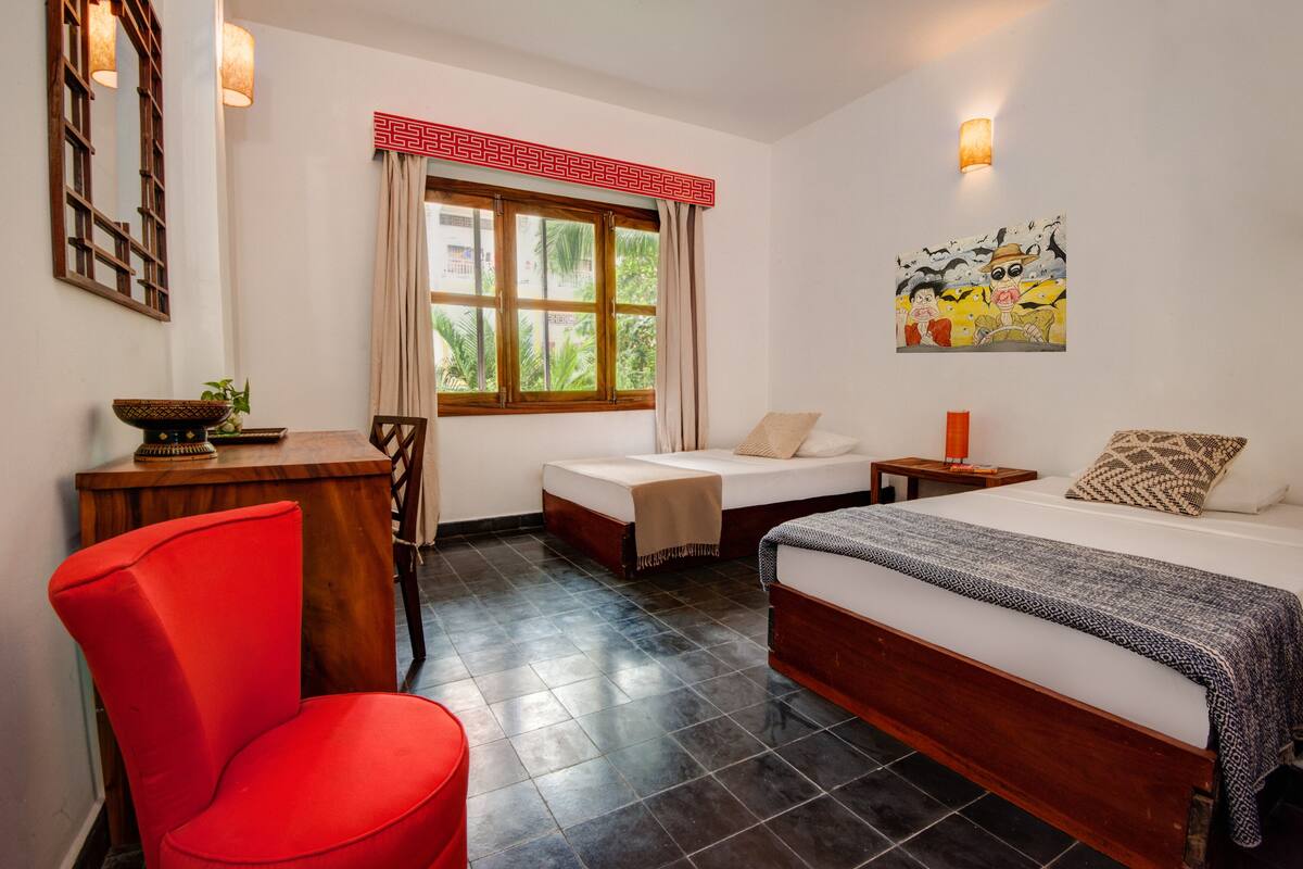 A hotel room with a bed, two bedside tables, a red chair, a wooden desk, a window with curtains, and a painting on the wall.