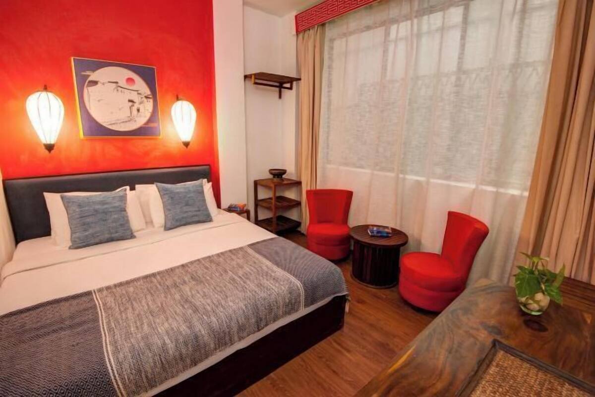 A hotel room with a bed, two red chairs, a small table, a window with curtains, and wall-mounted lights.