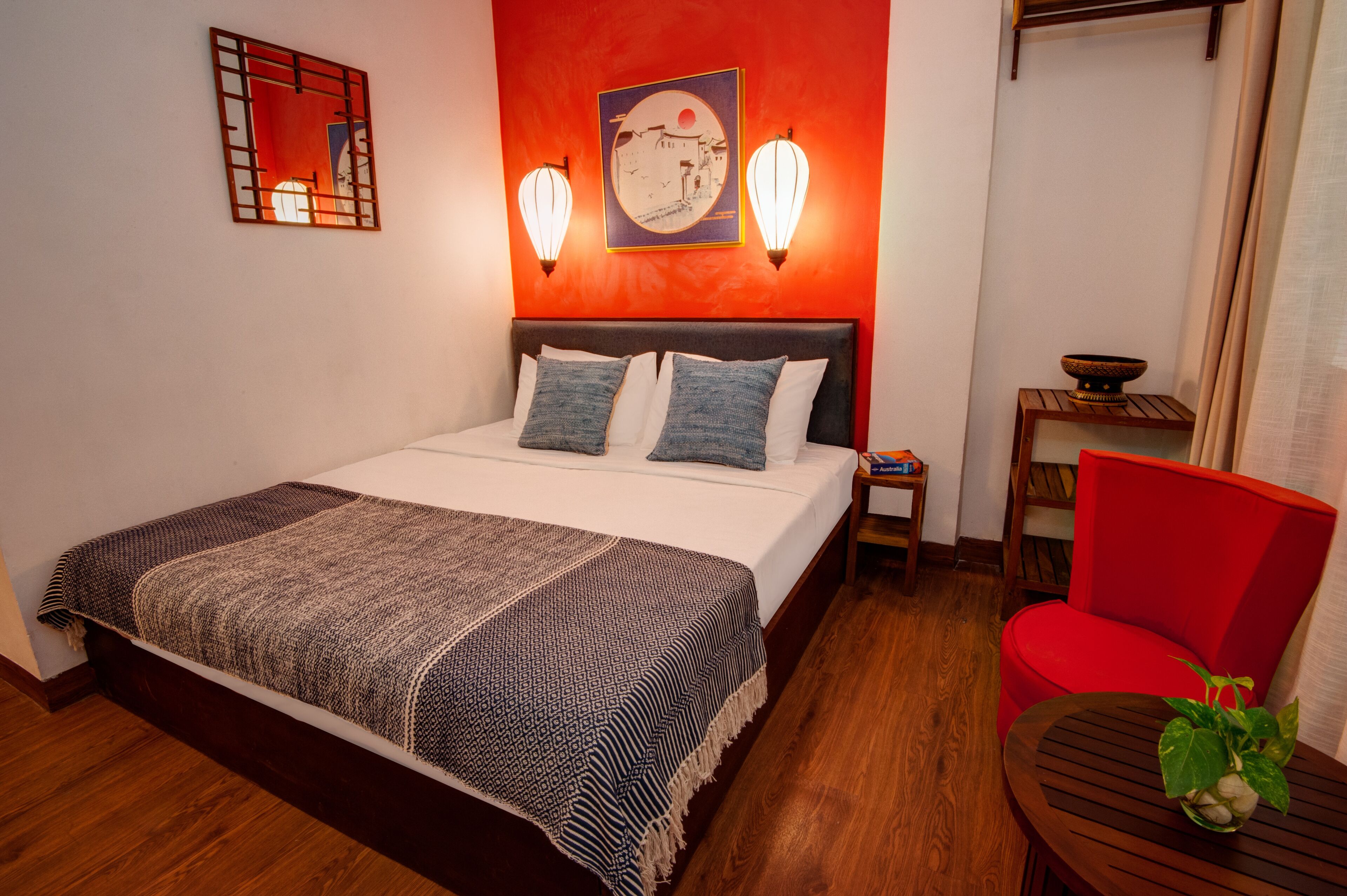 Habitación de hotel con cama, una silla roja, una mesita con un cuenco y una planta, y luces fijadas a la pared.