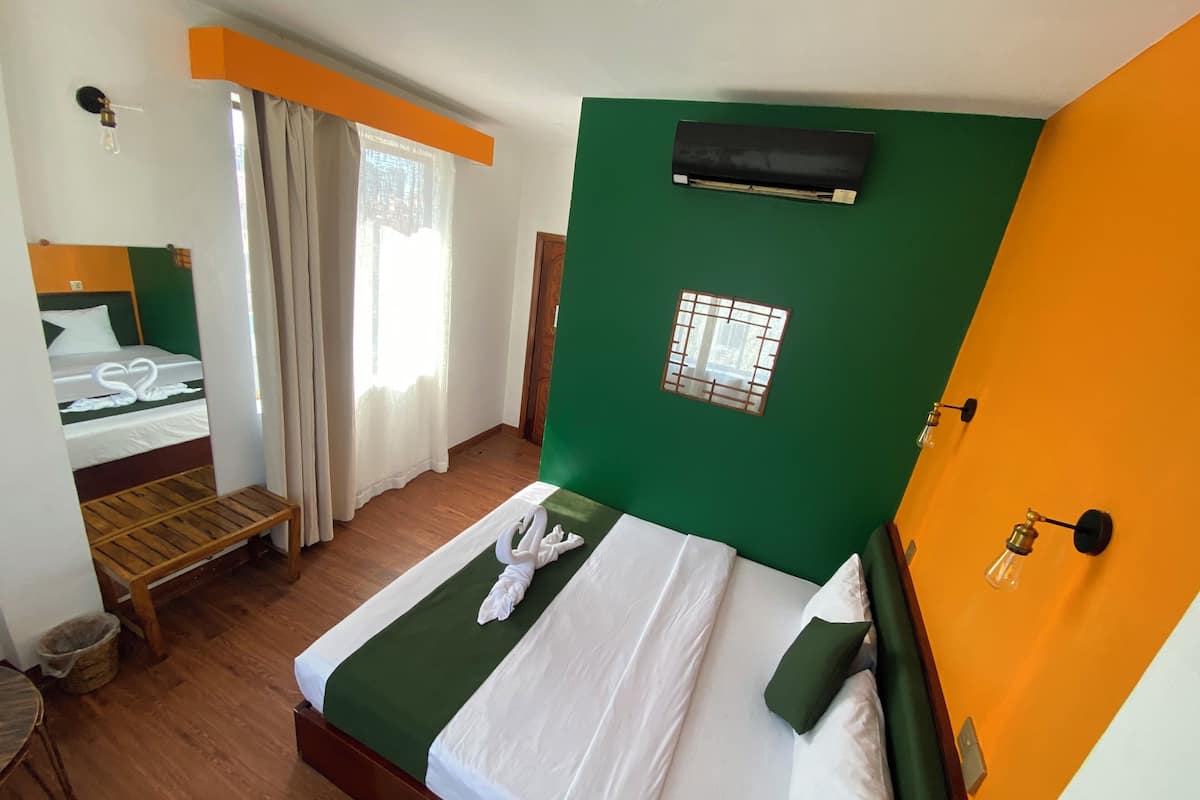 A hotel room with a bed, a small table, a chair, a window with curtains, and a wall-mounted air conditioner.