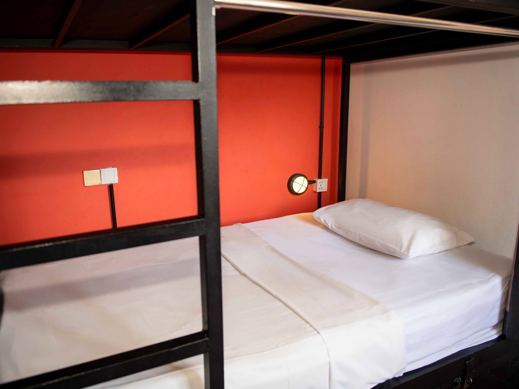 Una cama litera con ropa de cama blanca, una pared roja y una mesita de noche con una lámpara.