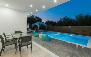 A modern outdoor pool area with a glass table and chairs, a covered seating area, and a wooden fence.