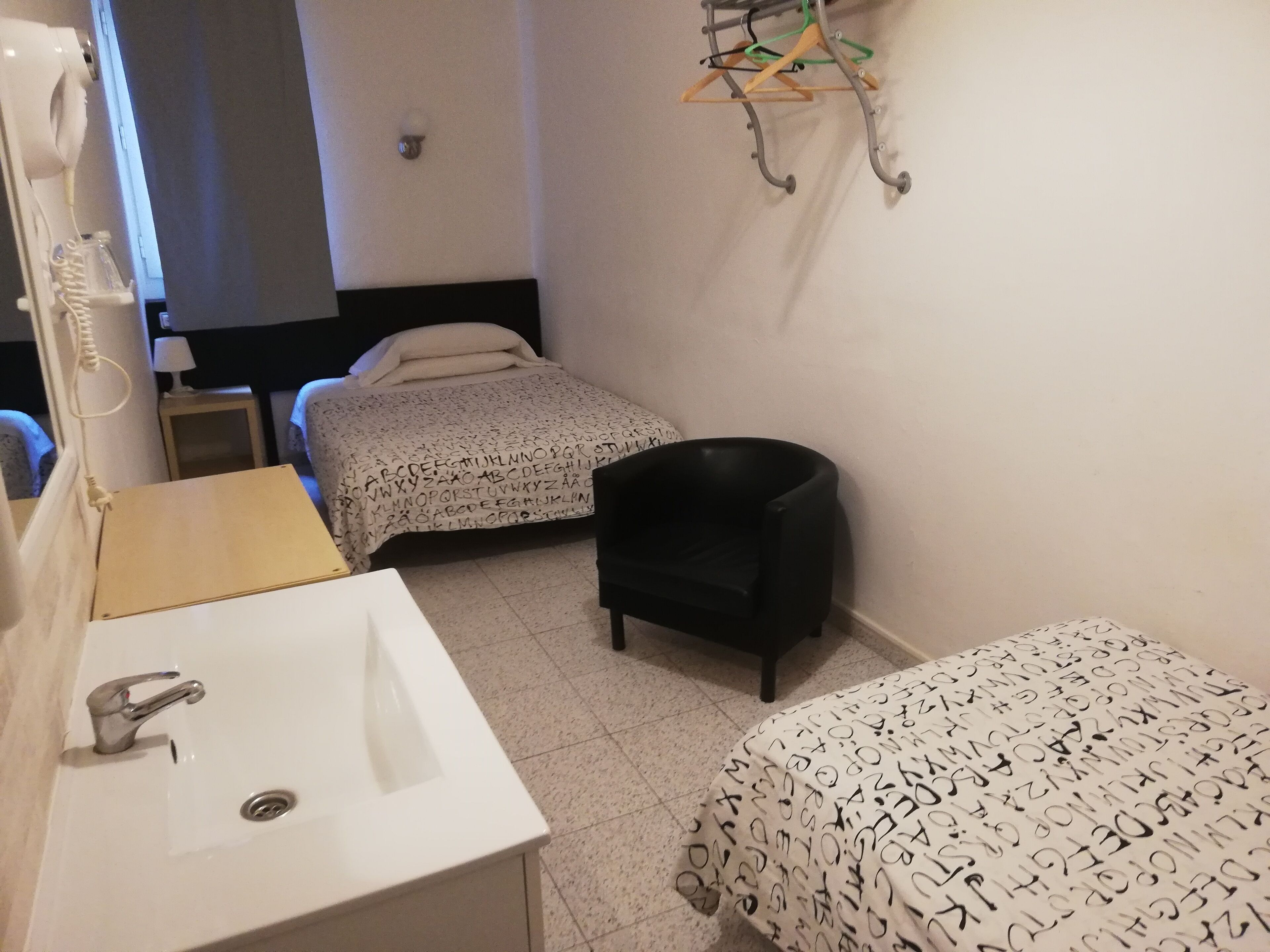 A small, clean room with a bed, a chair, a bedside table, and a bathroom with a sink and mirror.