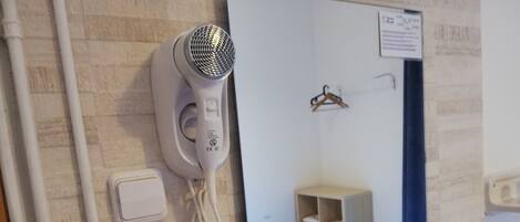 A hairdryer mounted on a wall with a mirror reflecting a room with a bed, a shelf, and a hanging coat rack.