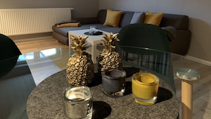 Uma sala de estar moderna com uma mesa de centro de vidro, dois abacaxis decorativos, uma vela e uma pequena mesa redonda com uma vela.