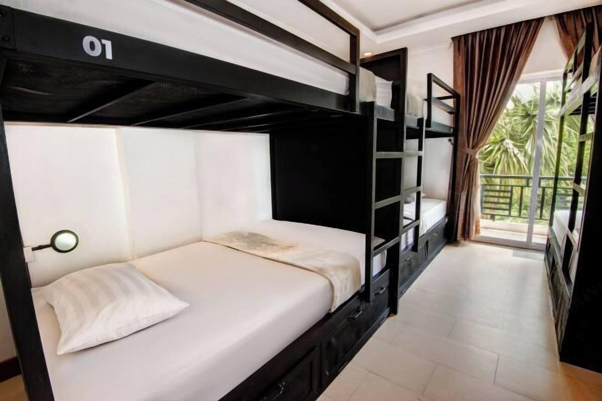 Una cama individual con el número '01' en la cabecera, una mesita de noche con lámpara y vistas al exterior a través de una ventana con cortinas.
