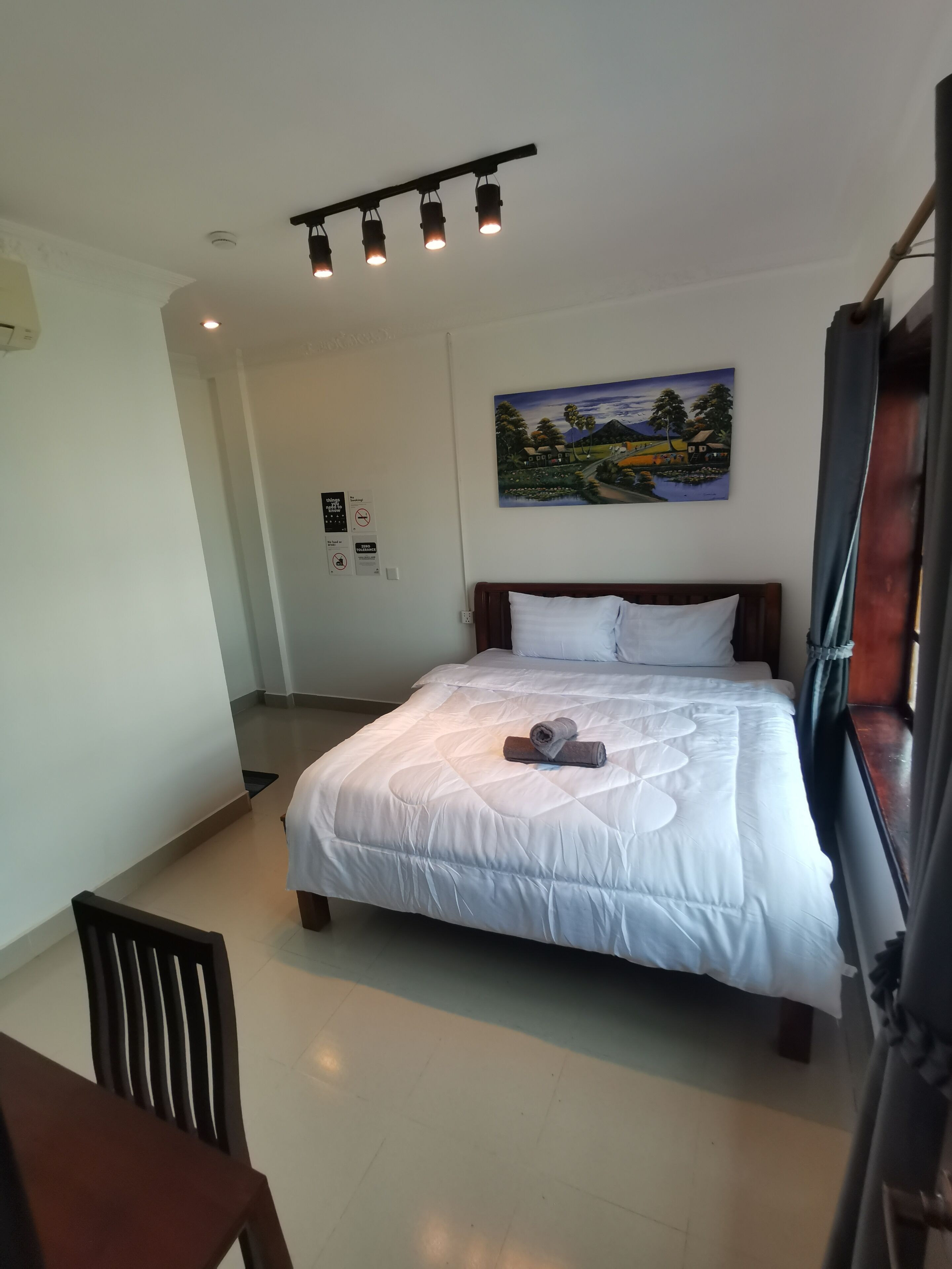 Una cama bien hecha con edredón blanco y almohadas, un cuadro de un paisaje en la pared y una lámpara en el techo.
