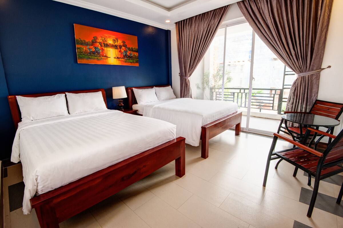 Habitación de hotel con dos camas, un cuadro en la pared, un balcón con cortinas y una mesa con sillas.