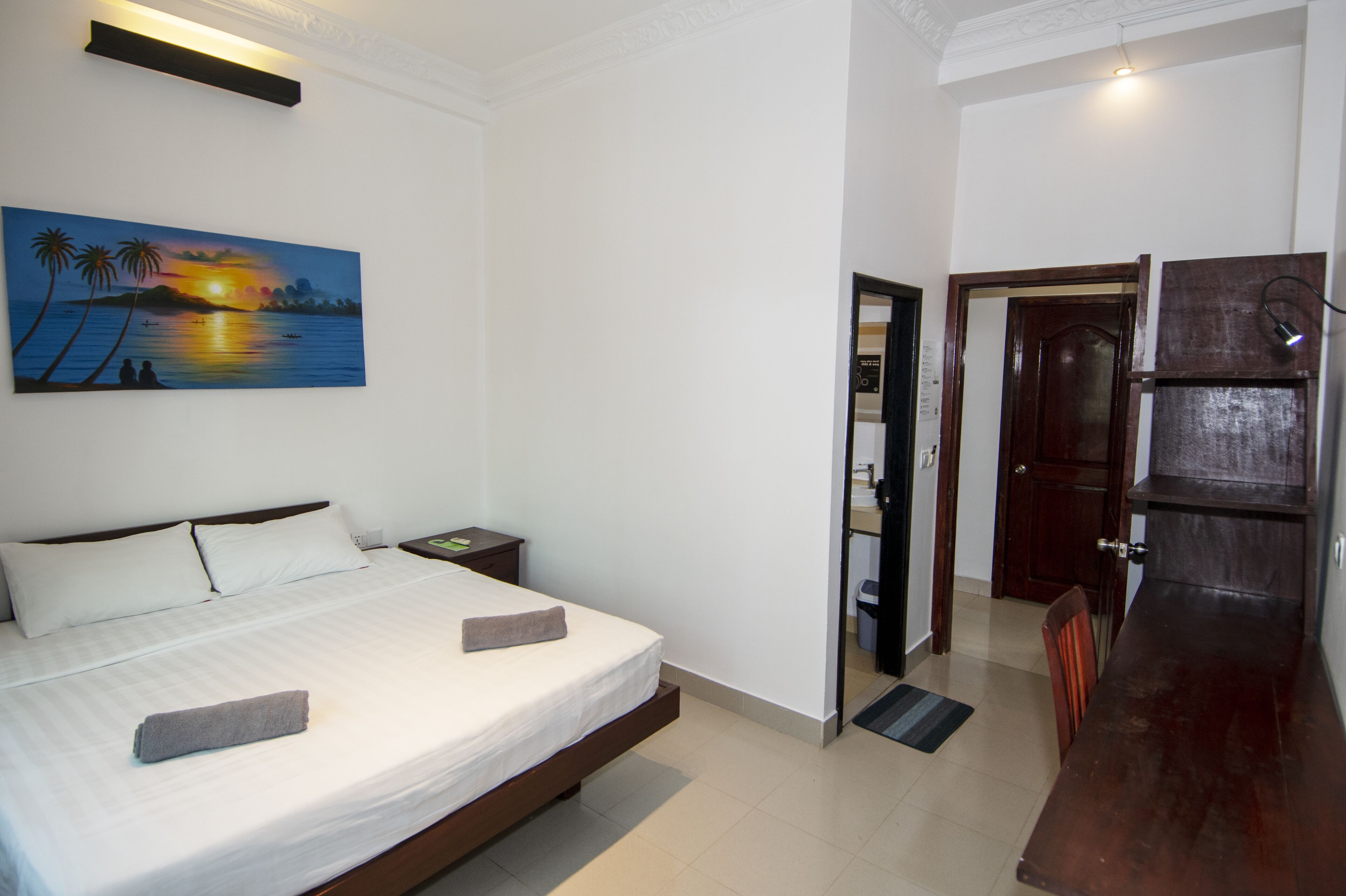 Una habitación de hotel con una cama, un cuadro de un atardecer, un banco de madera y una puerta que da a un baño.