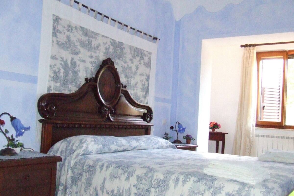 Un dormitorio con cama de madera, papel tapiz floral, una ventana con cortinas y una mesita de noche con lámpara.