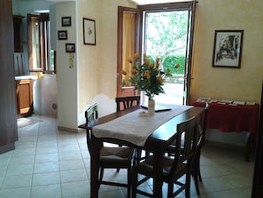 Un comedor con una mesa de madera, sillas y un jarrón de flores. Se observa una ventana con cortinas, una puerta y una pared decorada con cuadros.