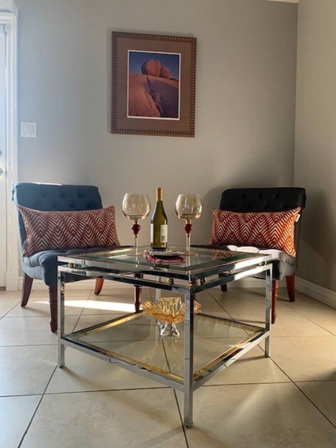 Una sala de estar con una mesa de centro de vidrio, dos sillas con cojines estampados, un cuadro enmarcado en la pared y una botella de vino.