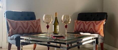 Una sala de estar con una mesa de centro de vidrio, dos sillas con cojines estampados, un cuadro enmarcado en la pared y una botella de vino.