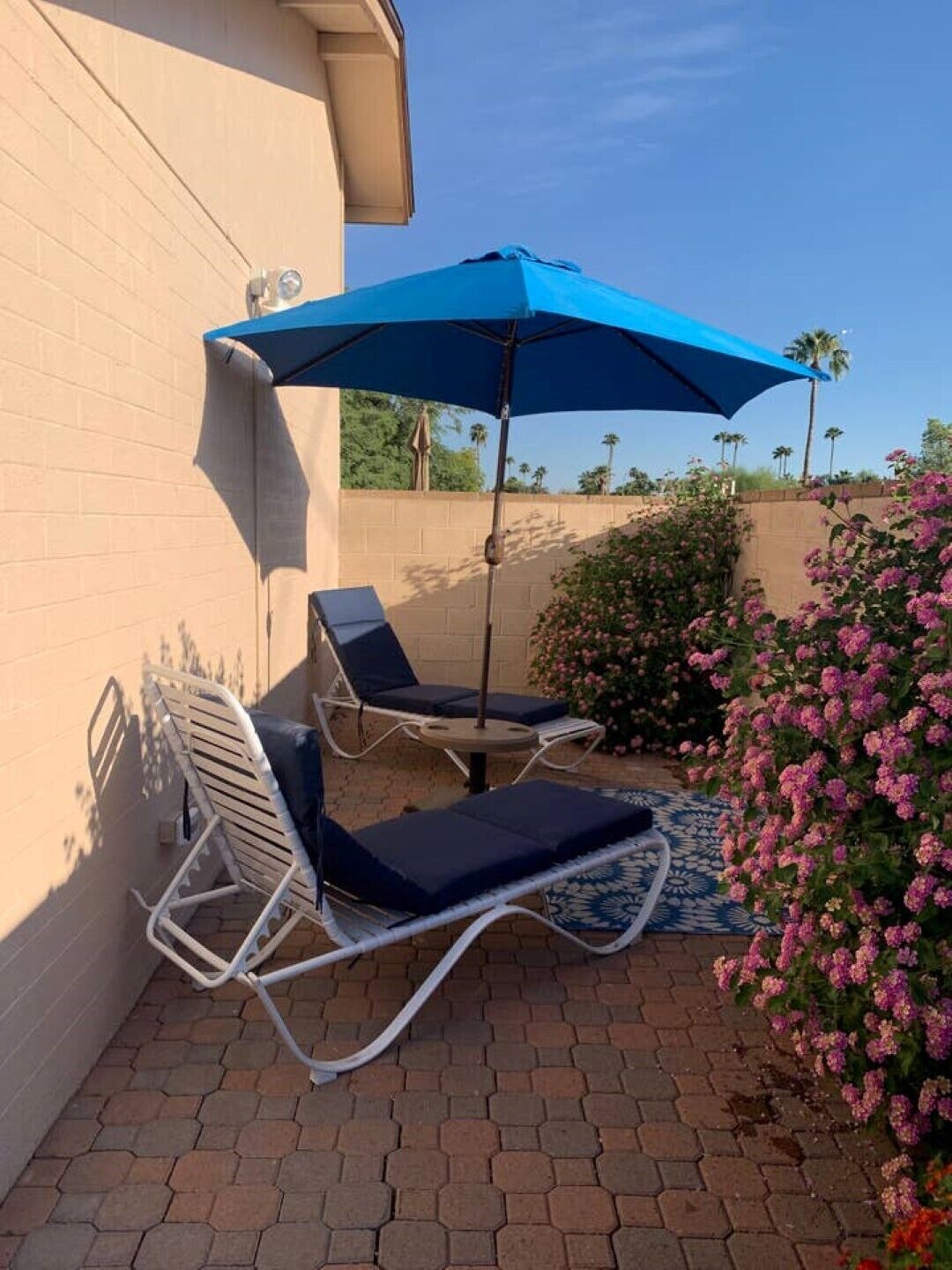 Un patio con dos sillones de descanso y una mesa bajo un paraguas azul, rodeado de pavimento de ladrillo y plantas con flores.