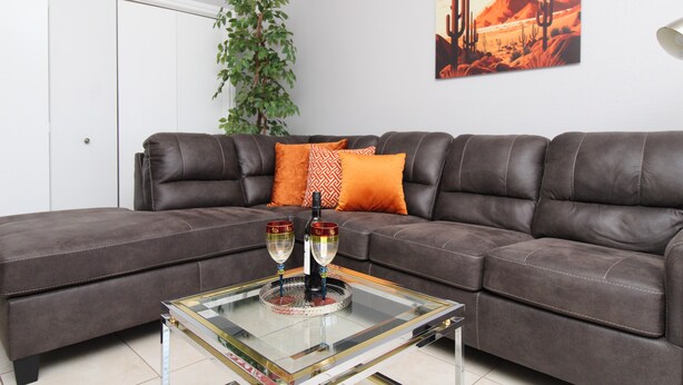 Un salon avec un canapé en cuir marron, deux coussins orange, une table basse en verre sur laquelle reposent deux verres à vin et une bouteille, un tableau représentant un cactus accroché au mur, et une plante verte.