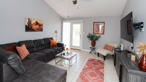 Un salon avec un canapé en cuir foncé, une table basse en verre, un tapis à motifs rouges, un ventilateur de plafond et un téléviseur à écran plat fixé au mur.