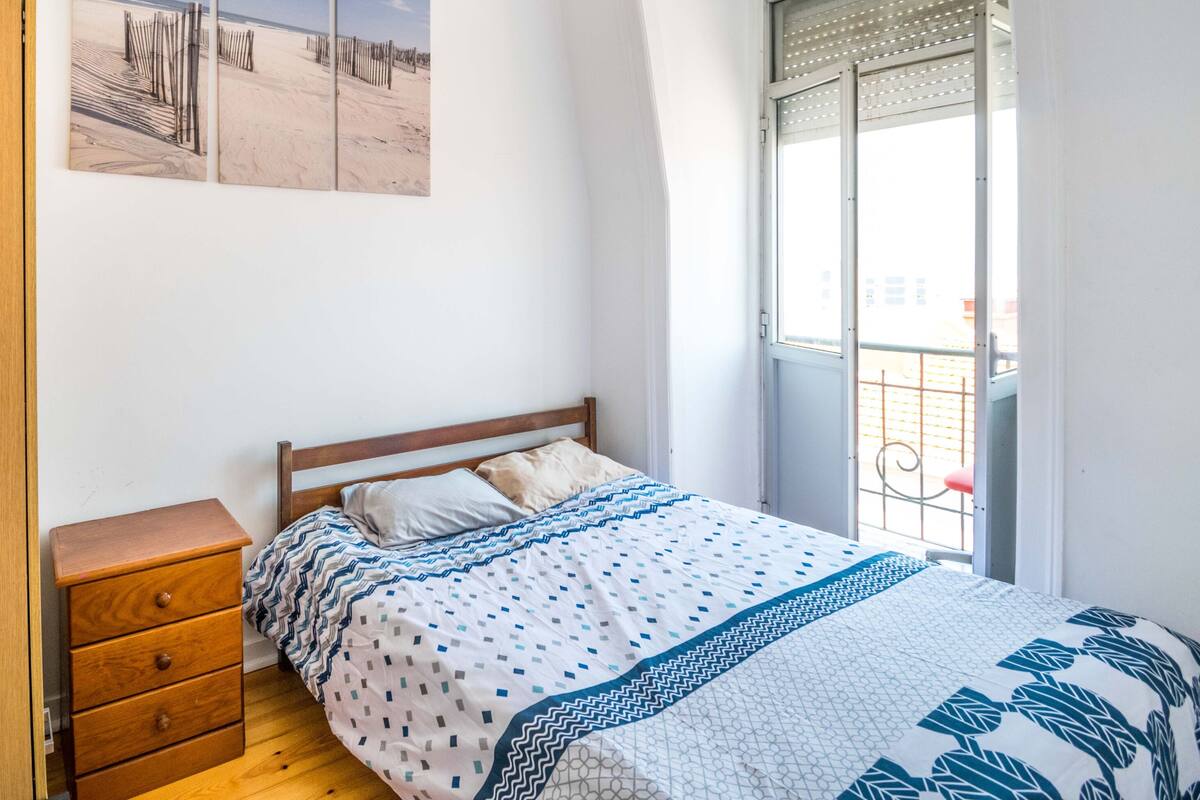 A bedroom with a wooden dresser, a bed with patterned bedding, a window with a view of the outside, and a wall with three framed pictures.