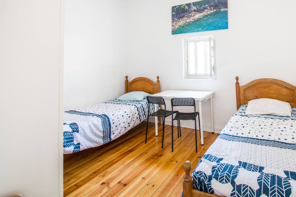 A room with two single beds, a small desk, a chair, and a window.