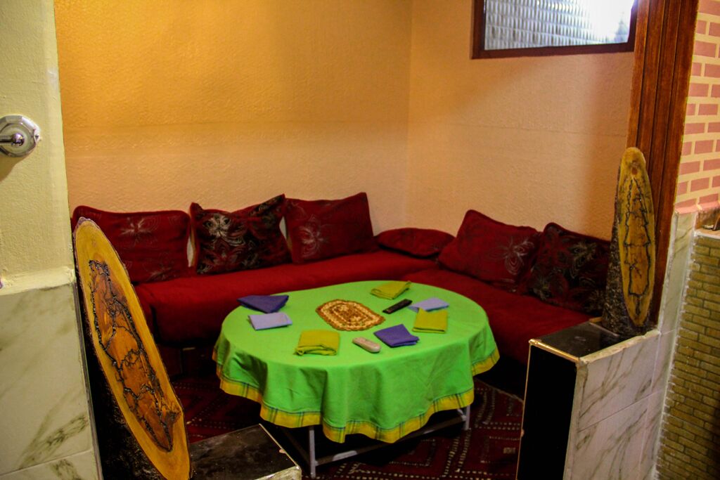 Ein kleiner Raum mit einem roten Sofa, einem runden Tisch mit grüner Tischdecke und einem hölzernen Gegenstand, der an der Wand lehnt.