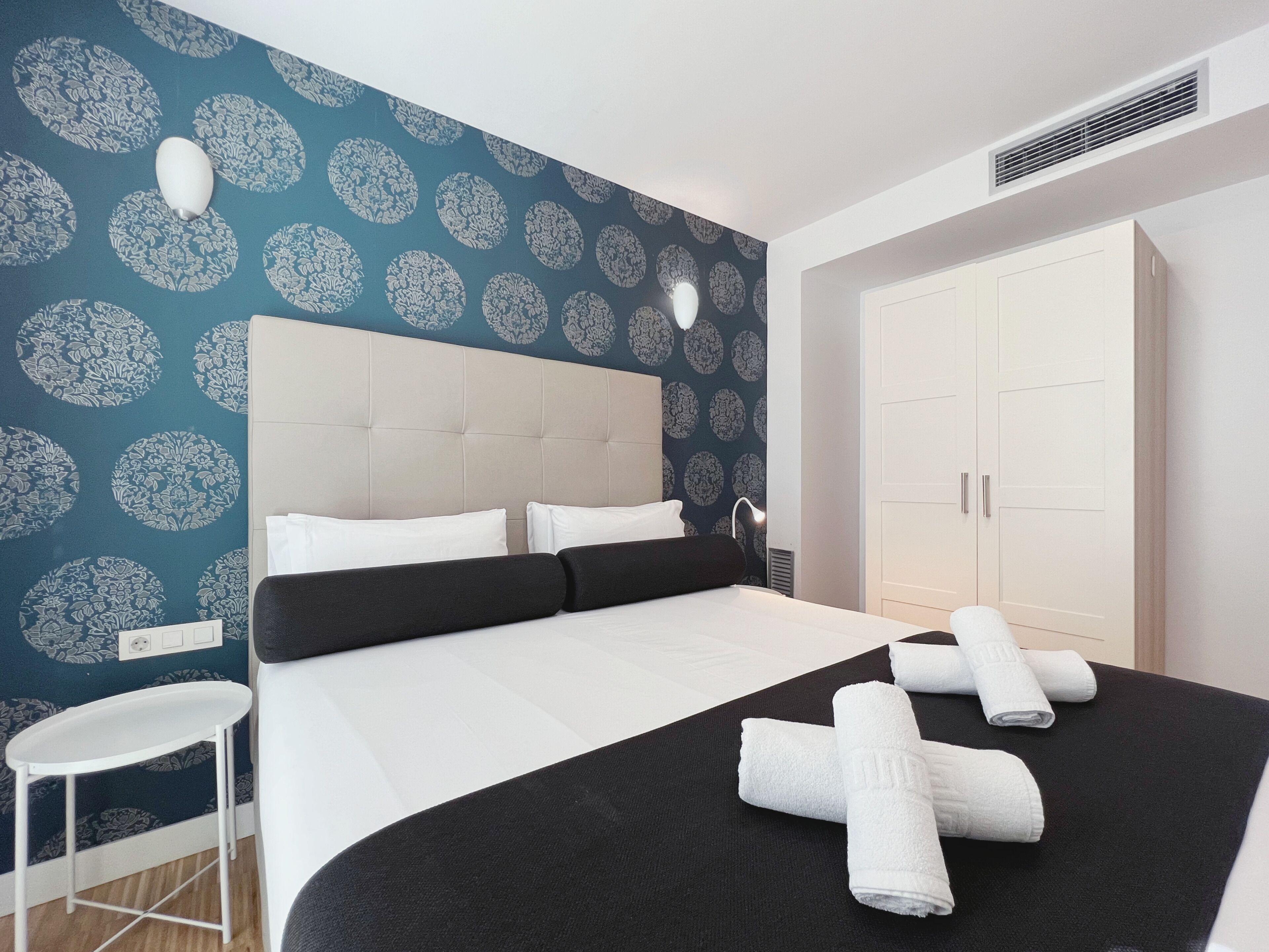 A hotel room with a bed, a white wardrobe, a small white table, and a patterned wall.