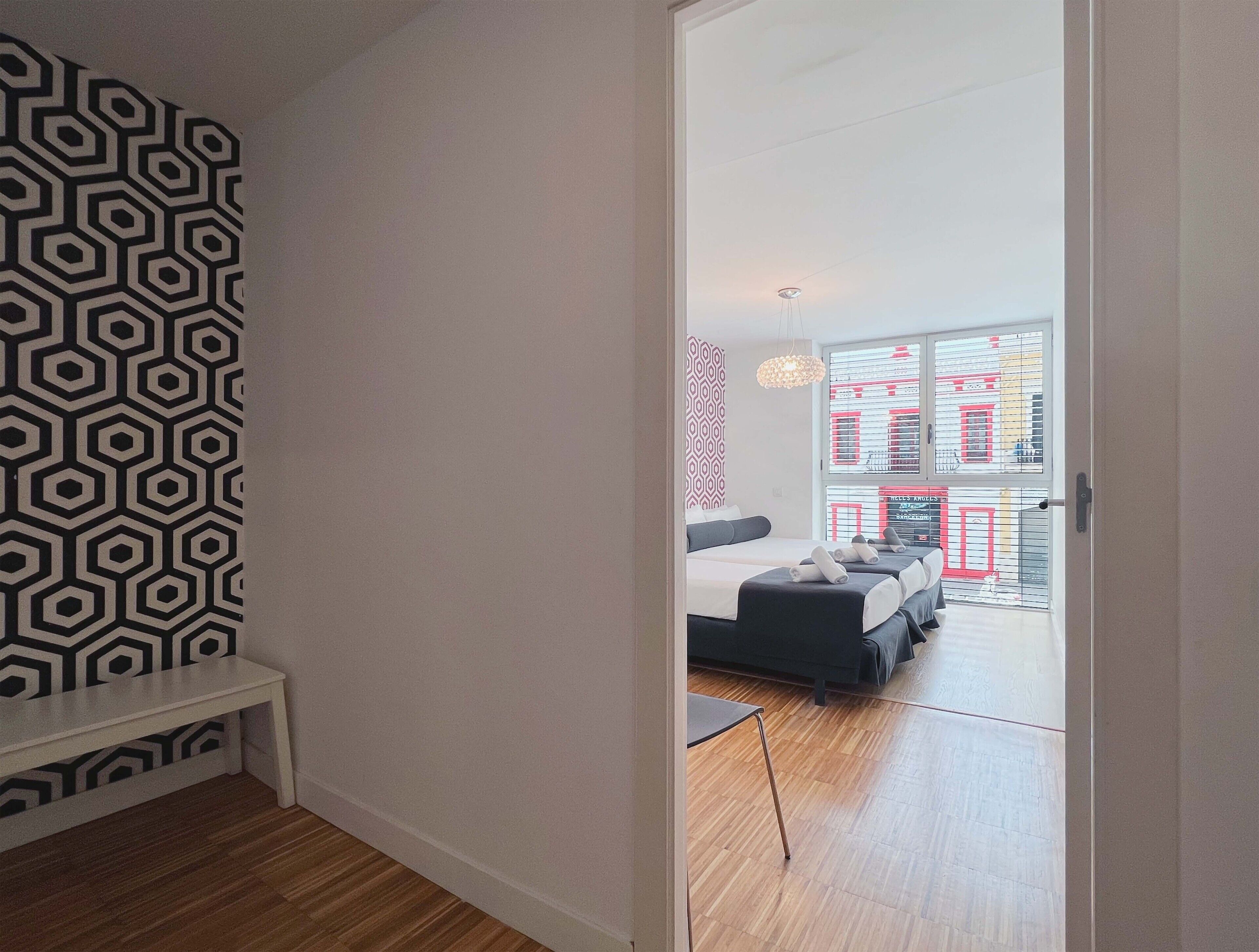 Une chambre d’hôtel moderne avec un mur orné d’un motif géométrique, un plancher en bois et une vue sur la ville grâce à de grandes fenêtres.