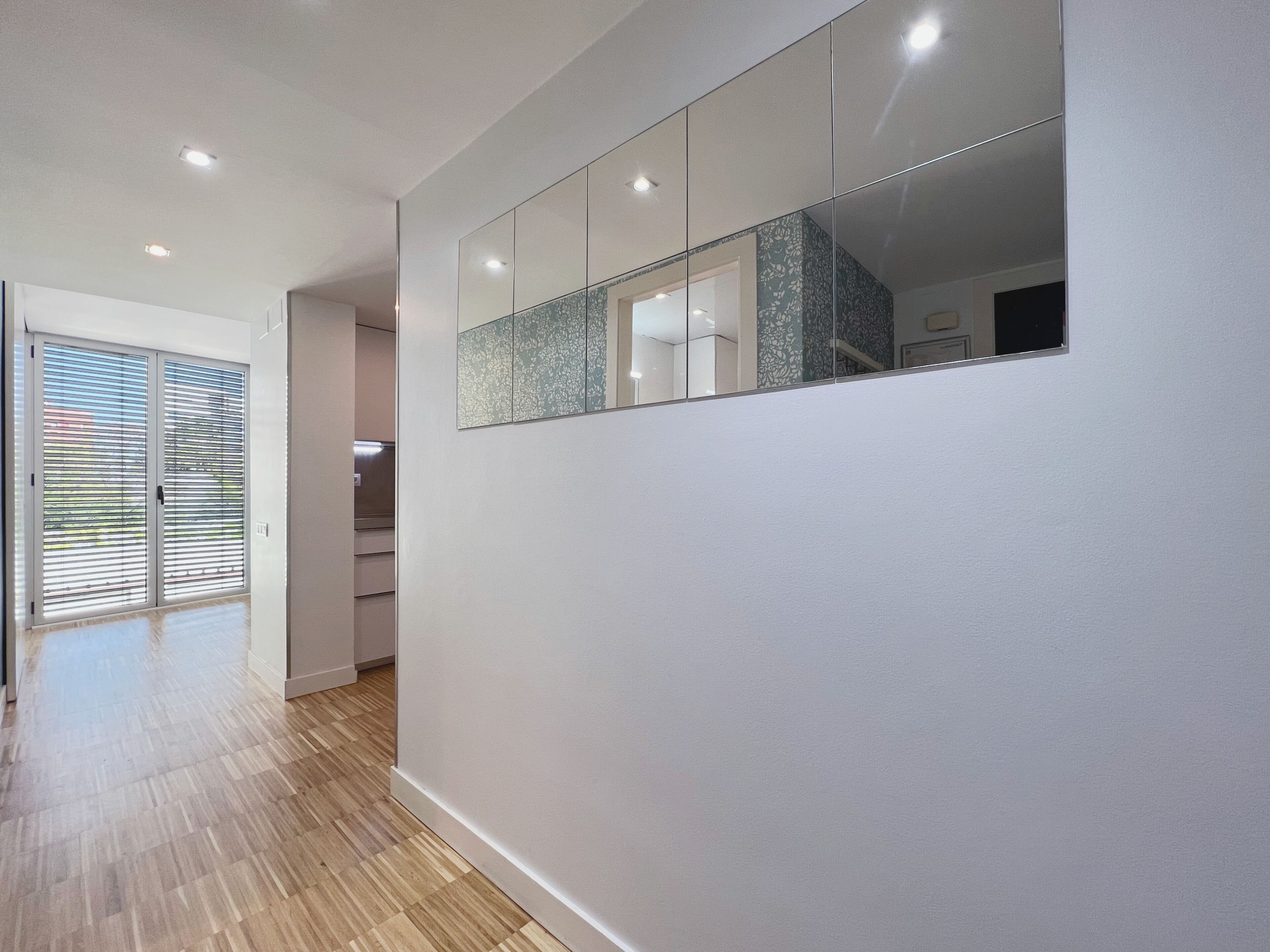 A hallway with a wooden floor, a sliding glass door, and a mirrored wall reflecting a kitchen area.