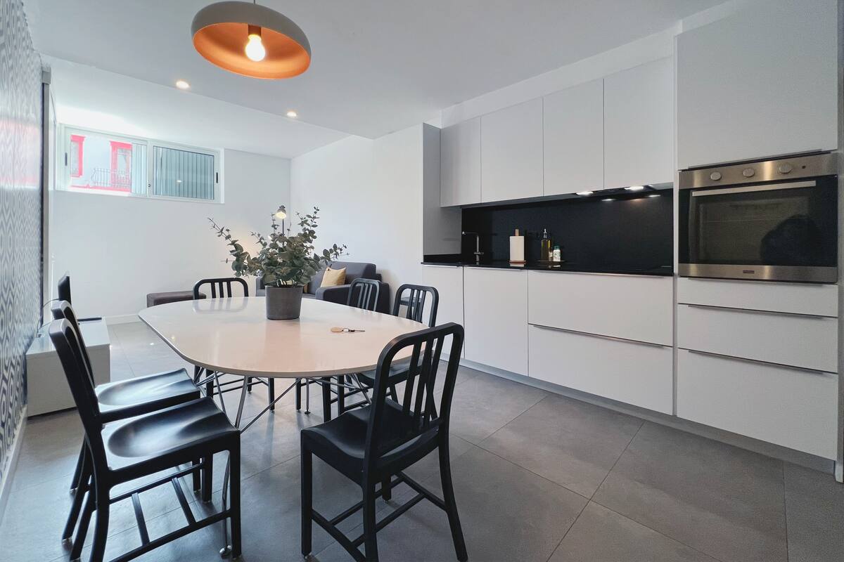 Una cocina moderna con gabinetes blancos, encimera negra y electrodomésticos de acero inoxidable. Hay una mesa de comedor con sillas y una planta en maceta.