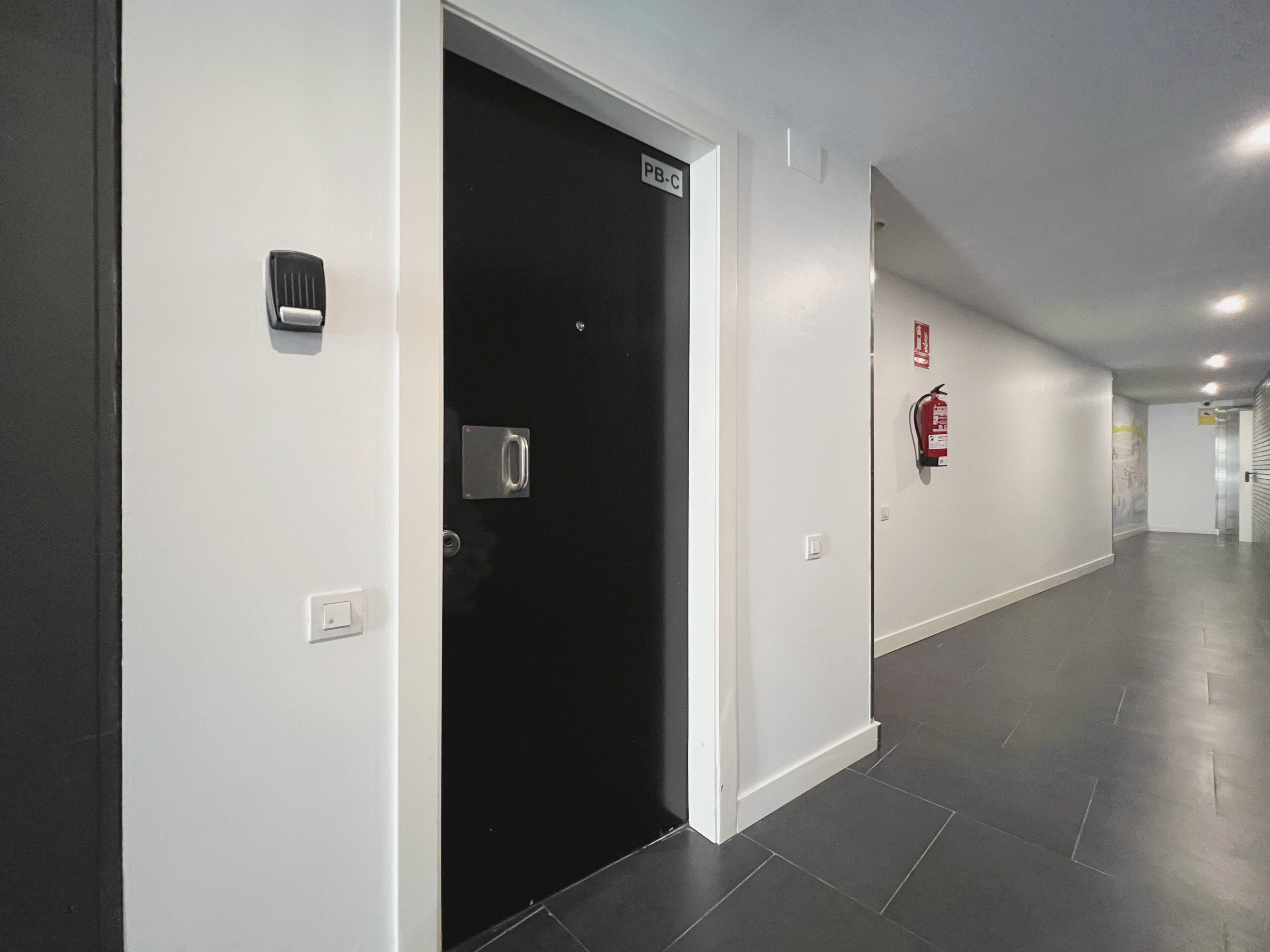 Un pasillo con una puerta negra identificada con la etiqueta 'PB-C', un extintor en la pared y un interruptor de luz.