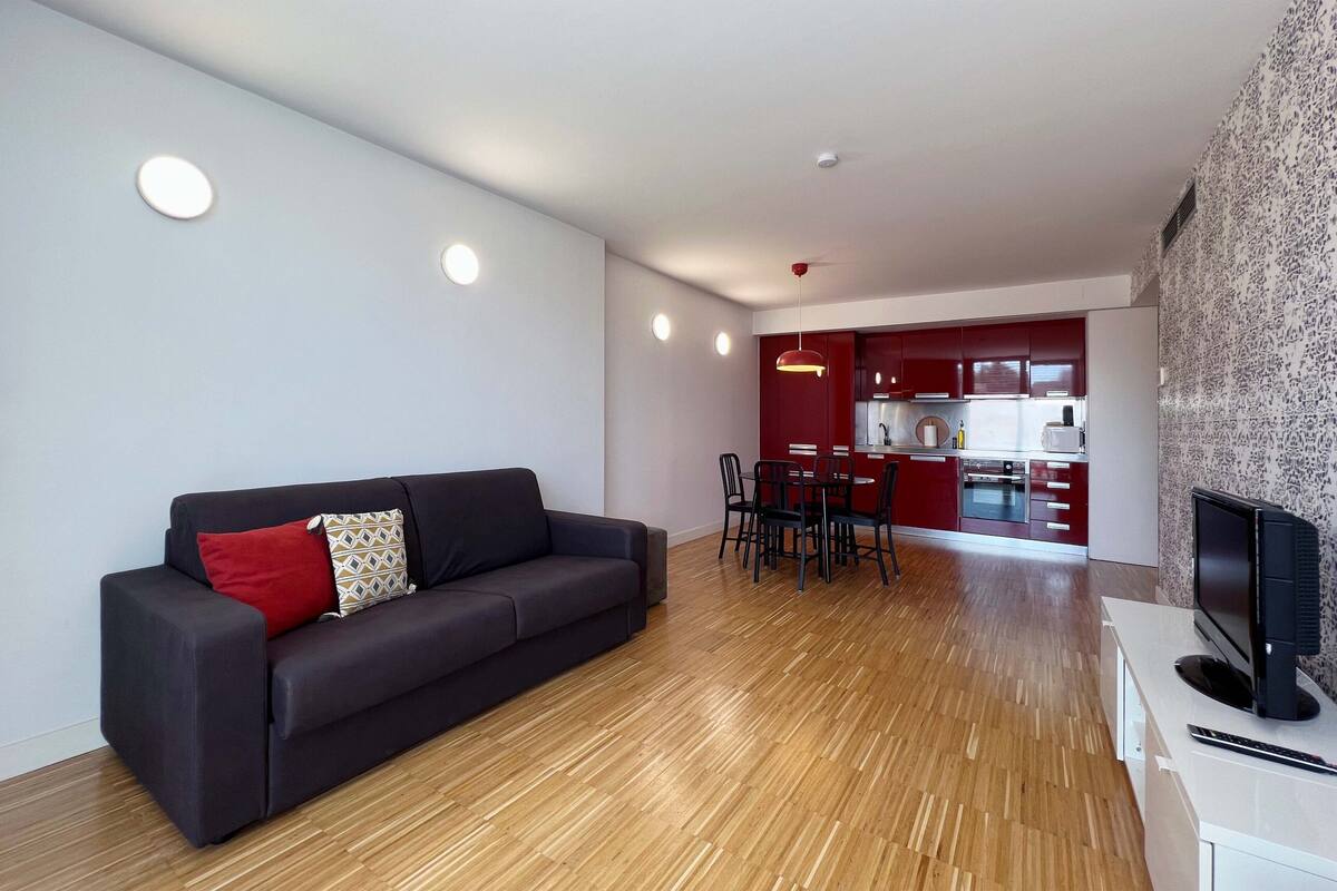Una sala de estar moderna con un sofá oscuro, un comedor con una mesa y sillas rojas, y una cocina al fondo.