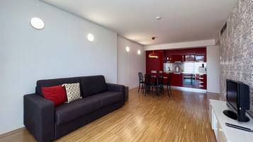 Een moderne woonkamer met een donkere bank, een eethoek met een rode tafel en stoelen, en een keuken op de achtergrond.