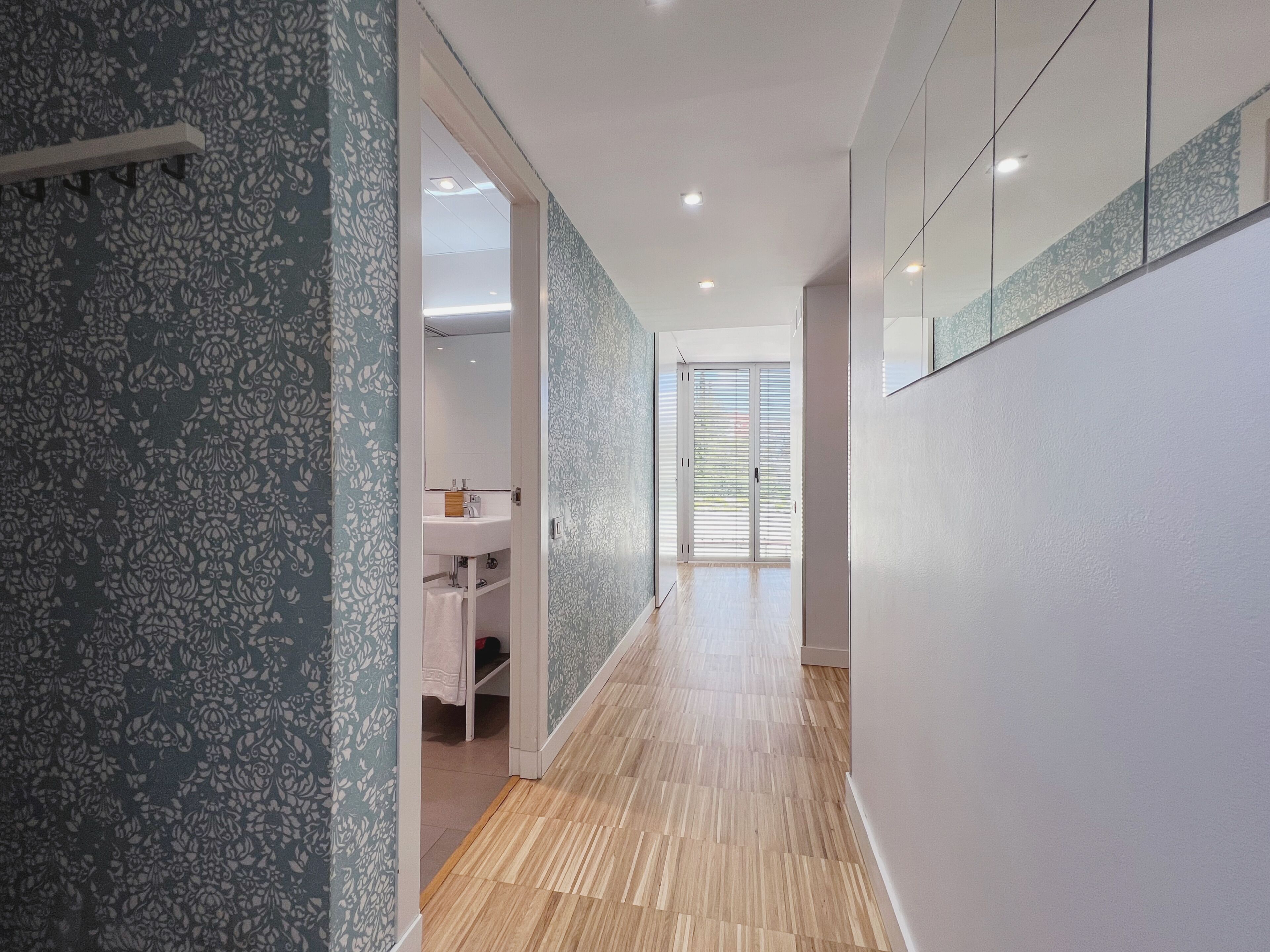 Ein Flur mit strukturiertem Wandpapier, Holzboden und Blick ins Badezimmer mit Waschbecken und Spiegel.