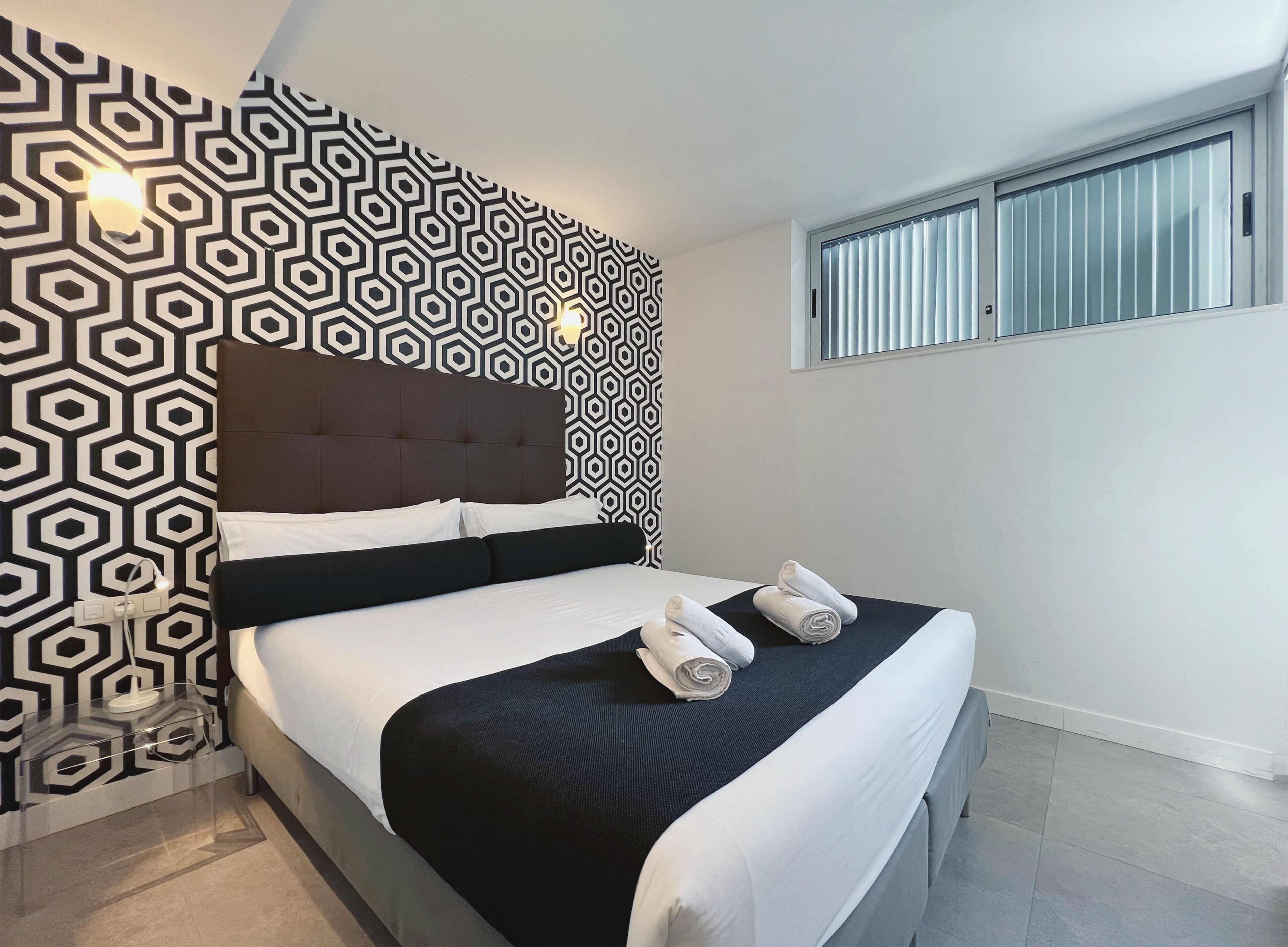 Una habitación de hotel moderna con una pared de diseño geométrico, una cama grande con ropa blanca y dos toallas enrolladas sobre la cama.