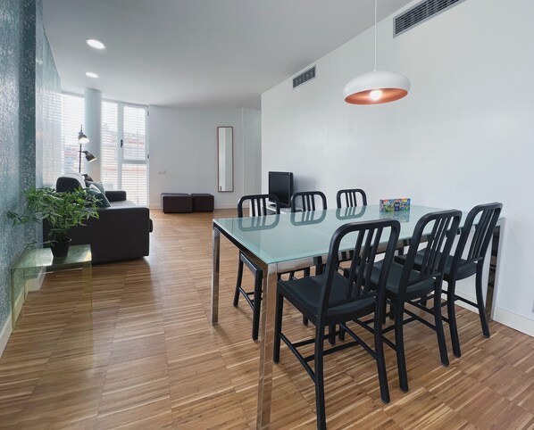 Un espace repas moderne avec une table en verre et des chaises noires, un canapé avec des coussins, une plante en pot et une grande fenêtre avec des stores.
