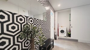 Una habitación moderna con un papel tapiz de diseño geométrico, un banco negro, una planta en maceta, una lavadora y una mopa.