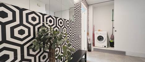 Una habitación moderna con un papel tapiz de diseño geométrico, un banco negro, una planta en maceta, una lavadora y una mopa.