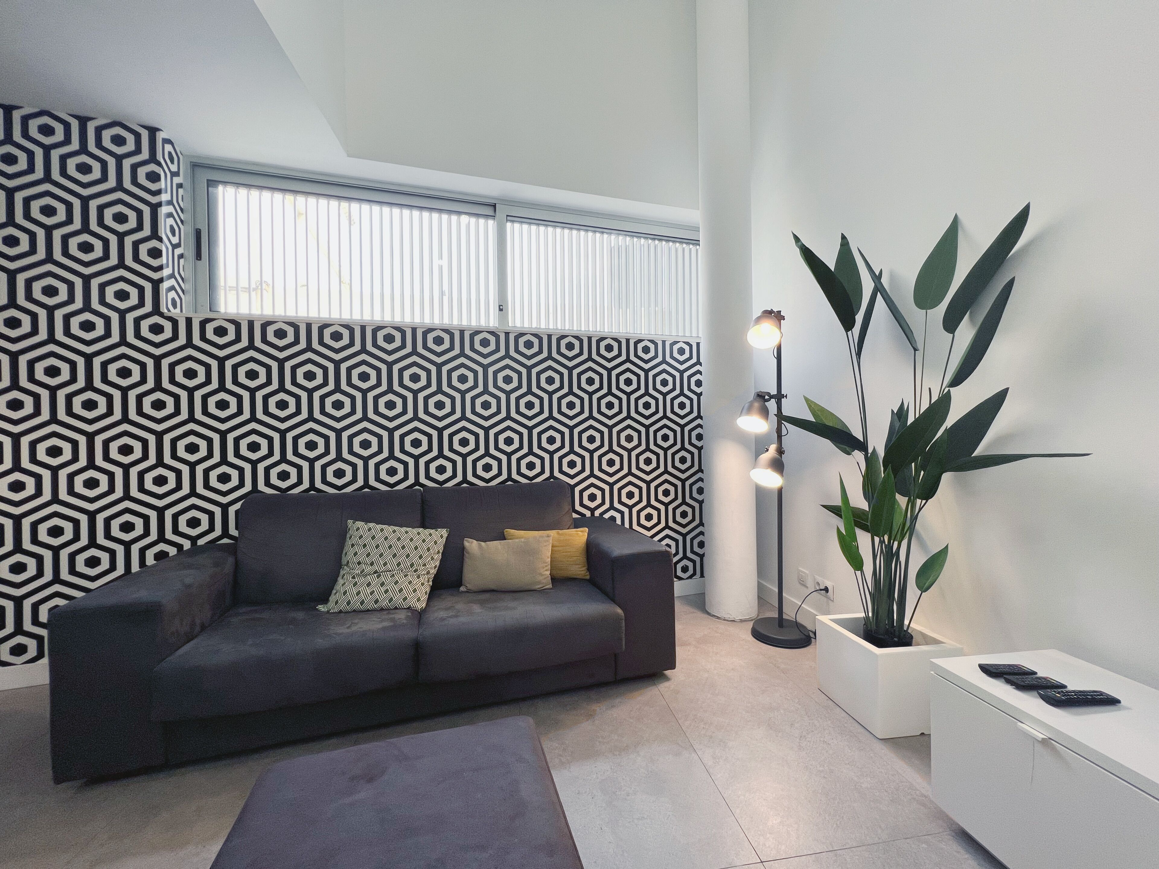 A modern living room with a dark grey sofa, patterned wallpaper, a large window, a floor lamp, and a potted plant.