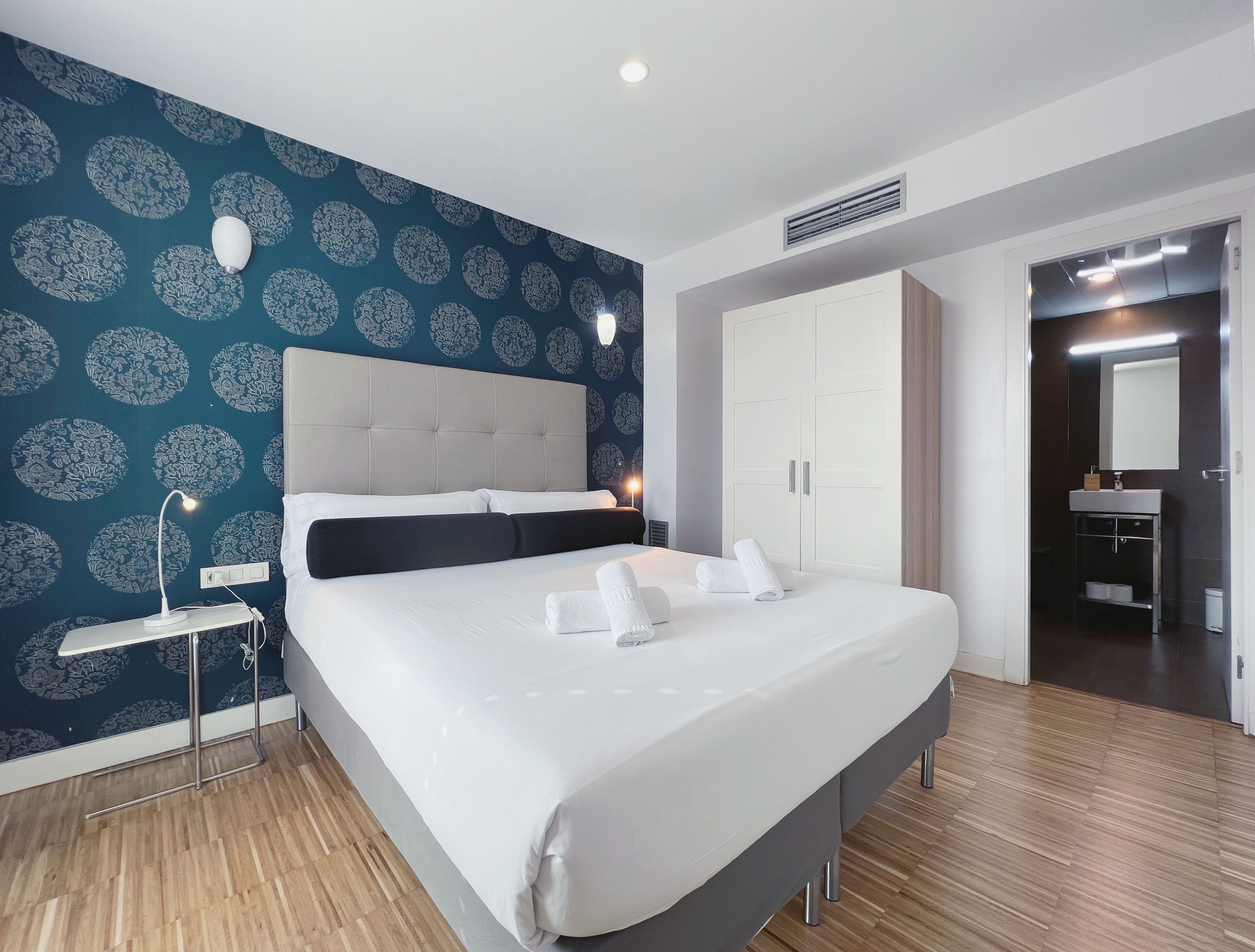 Une chambre d’hôtel moderne avec un grand lit, une table de chevet avec une lampe, une armoire et une salle de bain visible à travers une porte ouverte.