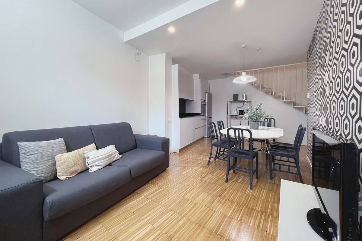 Una sala de estar moderna con un sofá gris, una mesa de comedor con sillas y una zona de cocina con microondas y estufa.