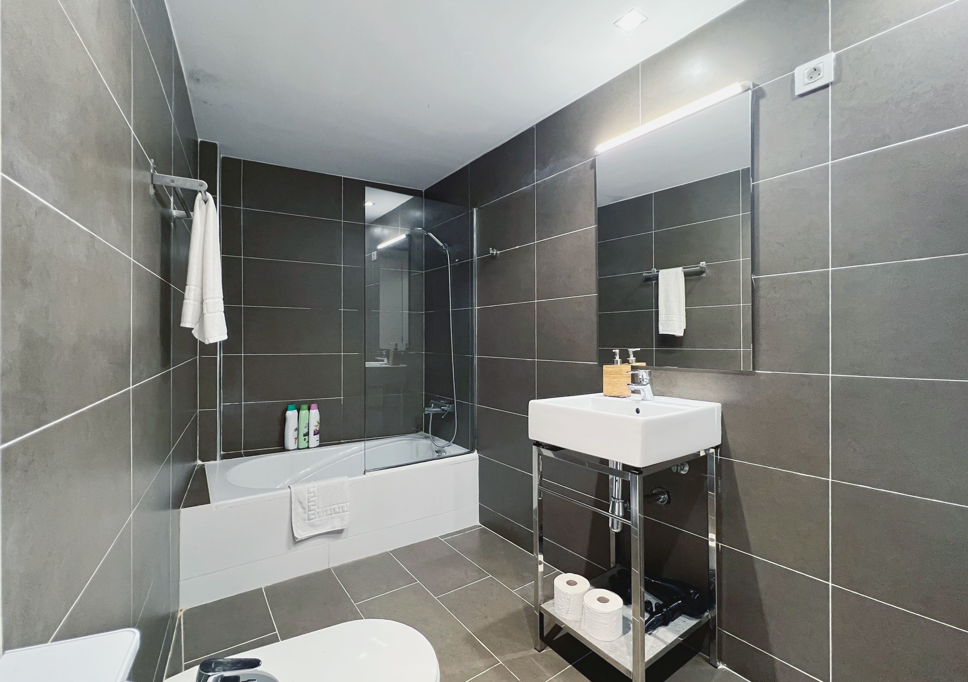 A modern bathroom with dark tiles, a white sink, a bathtub, and a glass shower enclosure.