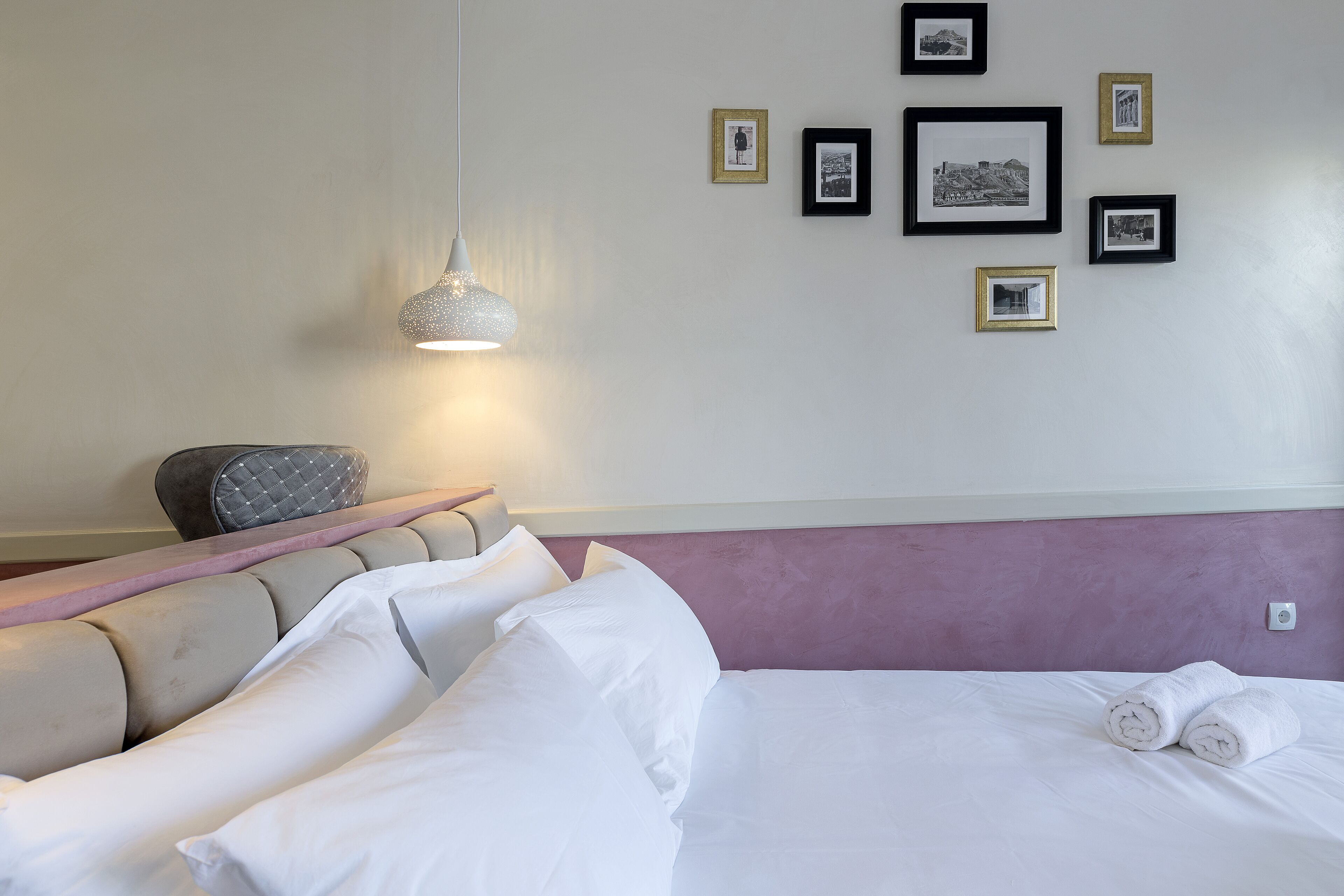 A hotel room with a bed, pillows, a headboard, a bedside table, a chair, and a wall with framed pictures.