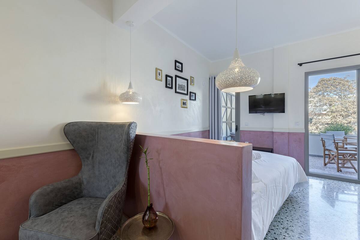 A modern hotel room with a large bed, a grey armchair, a small table with a vase, and a TV mounted on the wall.
