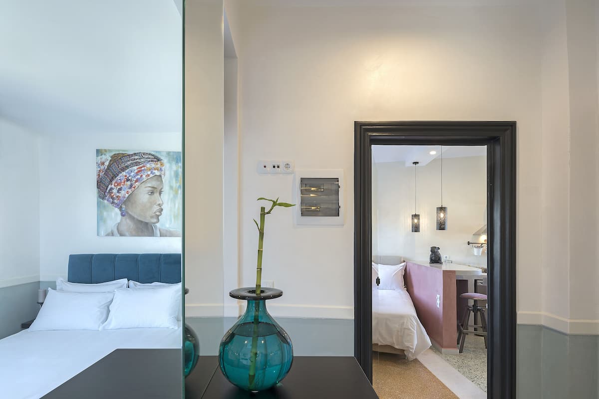 A modern hotel room with a bed, a vase with a plant, a painting on the wall, and a bathroom visible through an open door.