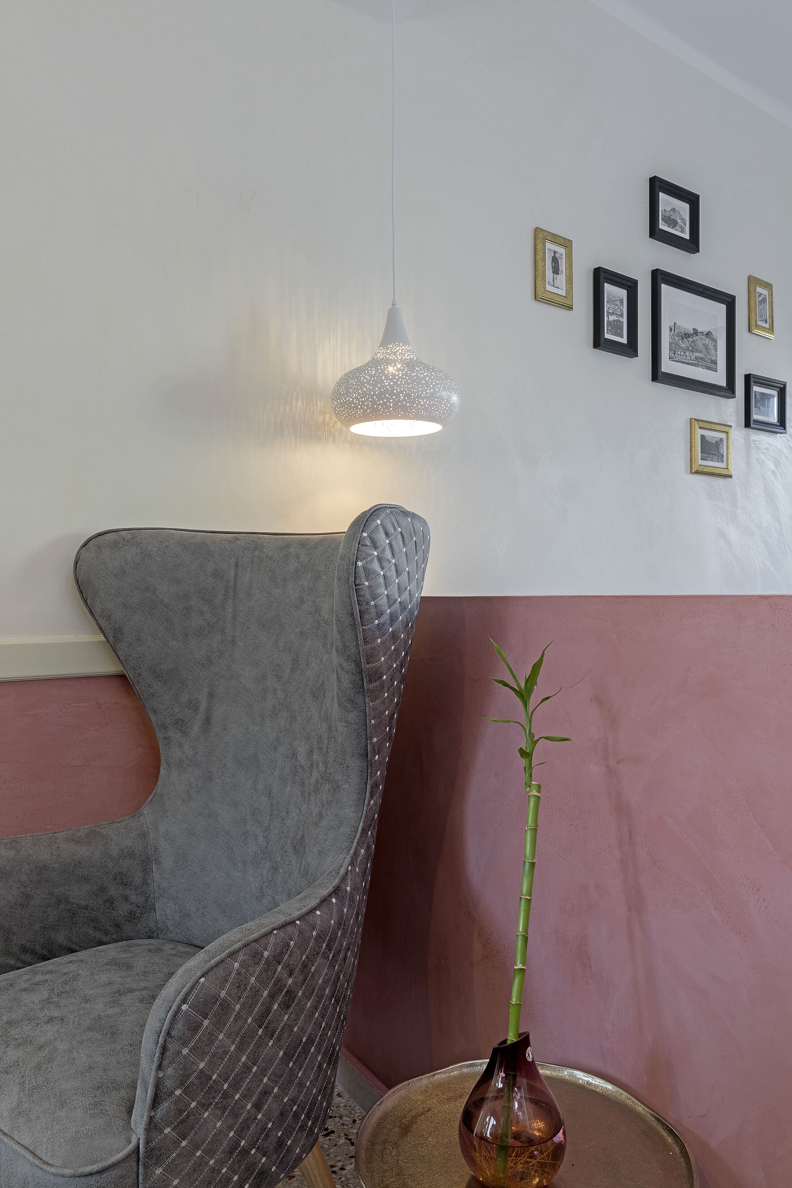 Ein Zimmer mit einem grau gepolsterten Sessel, einem kleinen runden Tisch mit einer Vase und einer Wand mit gerahmten Bildern.