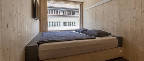 Ein kleines Schlafzimmer mit Holzwänden, einem Bett, einem Fenster und einem an der Wand befestigten Fernseher.