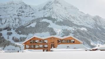 A wooden chalet with multiple windows, set against a backdrop of snow-covered mountains.