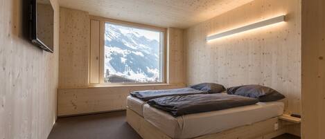 Een houten kamer met een bed, een raam met uitzicht op de bergen en een aan de muur bevestigde televisie.