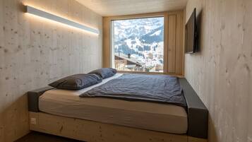 Une chambre avec un plafond en lambris de bois, un lit avec un matelas et des oreillers, une fenêtre donnant sur des montagnes enneigées et un téléviseur à écran plat fixé au mur.