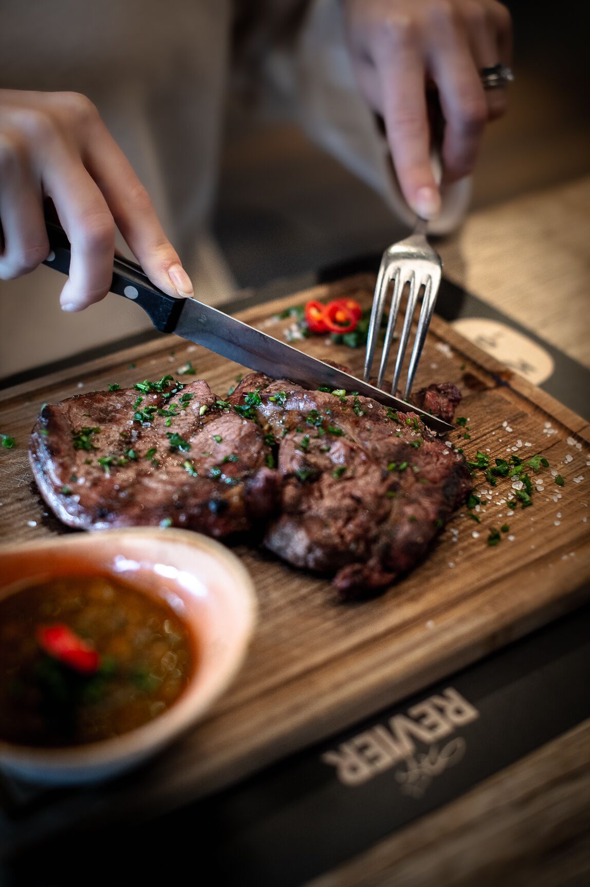 Una persona cortando un filete sobre una tabla de madera con un tenedor y un cuchillo.