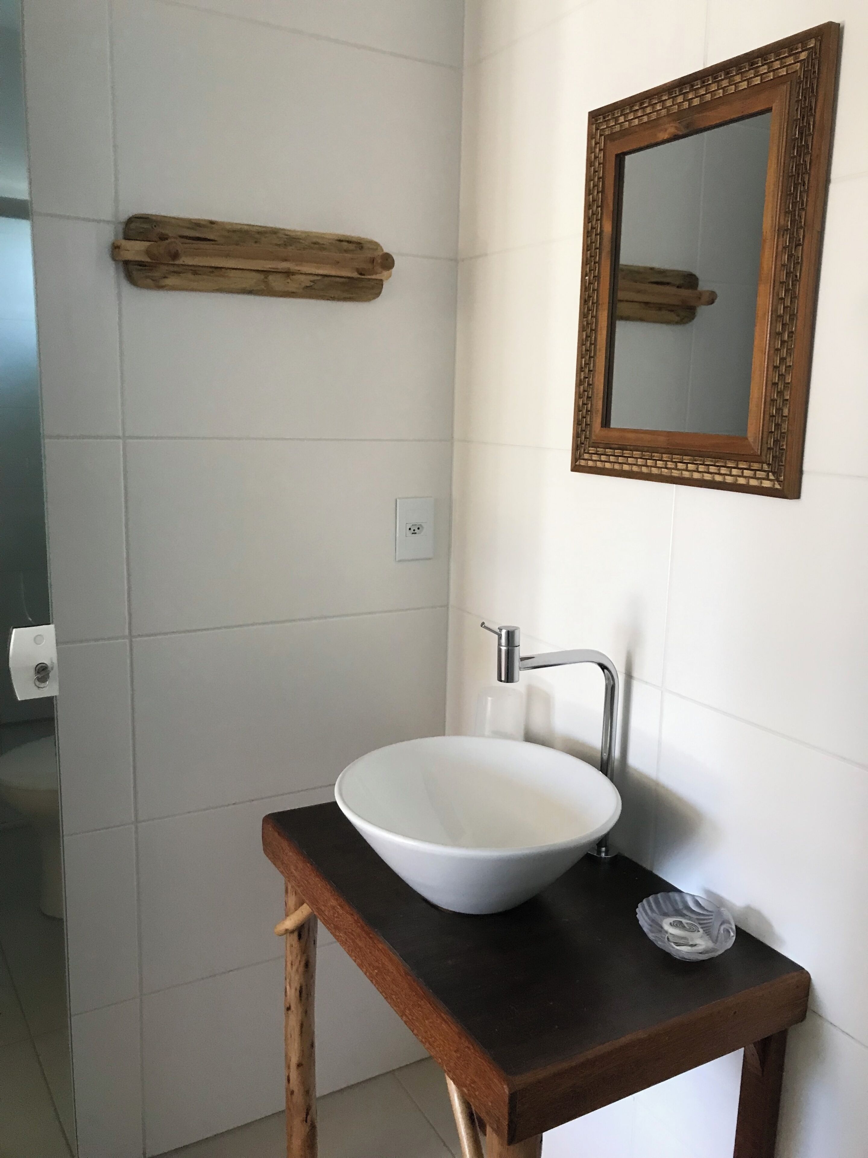 Um banheiro com parede de azulejos brancos, prateleira de madeira, pia branca com torneira moderna e espelho com moldura de madeira.